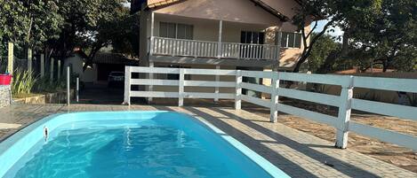A swimming pool with a white fence, a house with a balcony, and trees in the background.