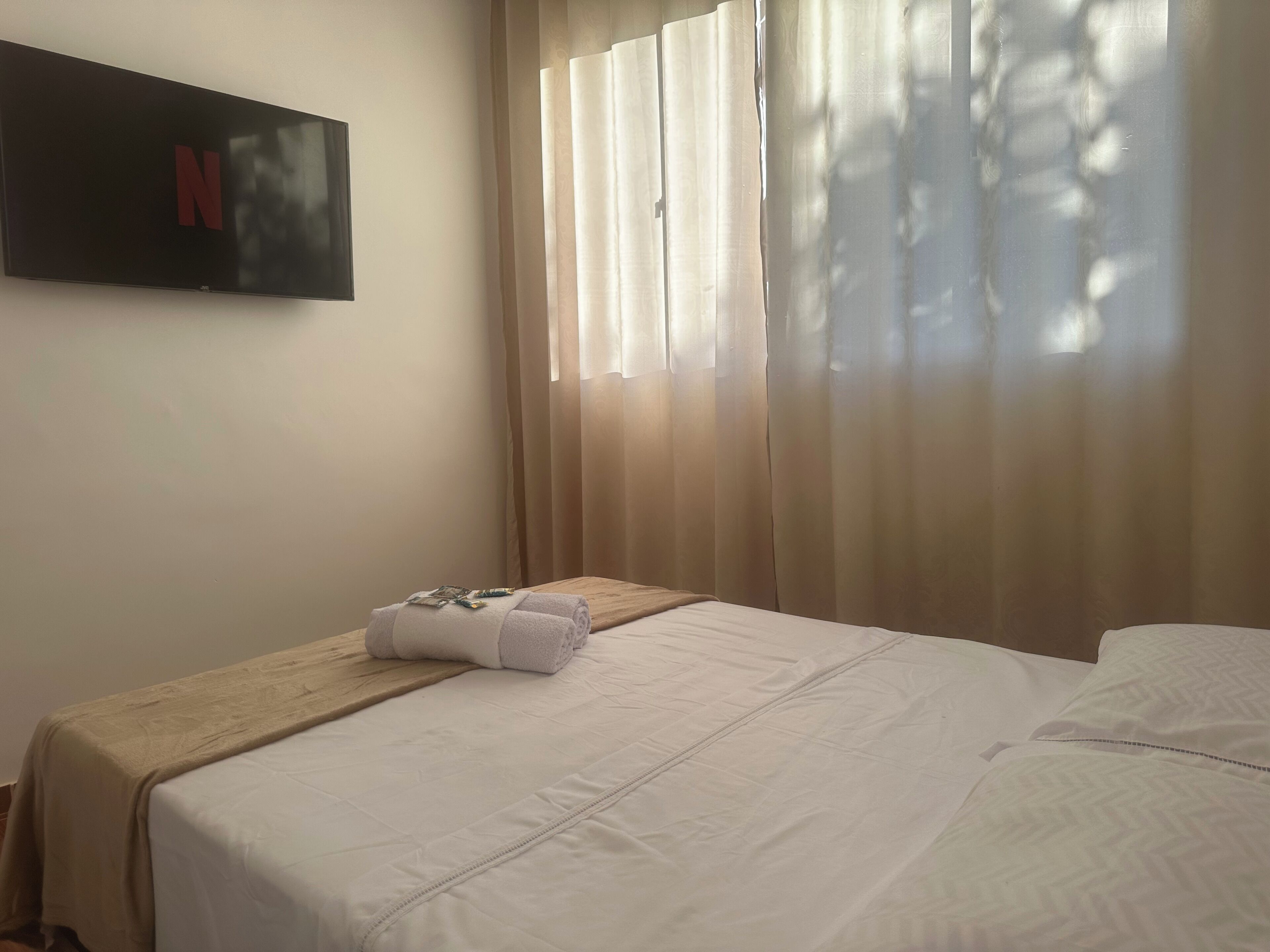Ein Hotelzimmer mit einem Bett, einem Nachttisch aus Holz, einem an der Wand montierten Fernseher und einem Fenster mit durchsichtigen Vorhängen.