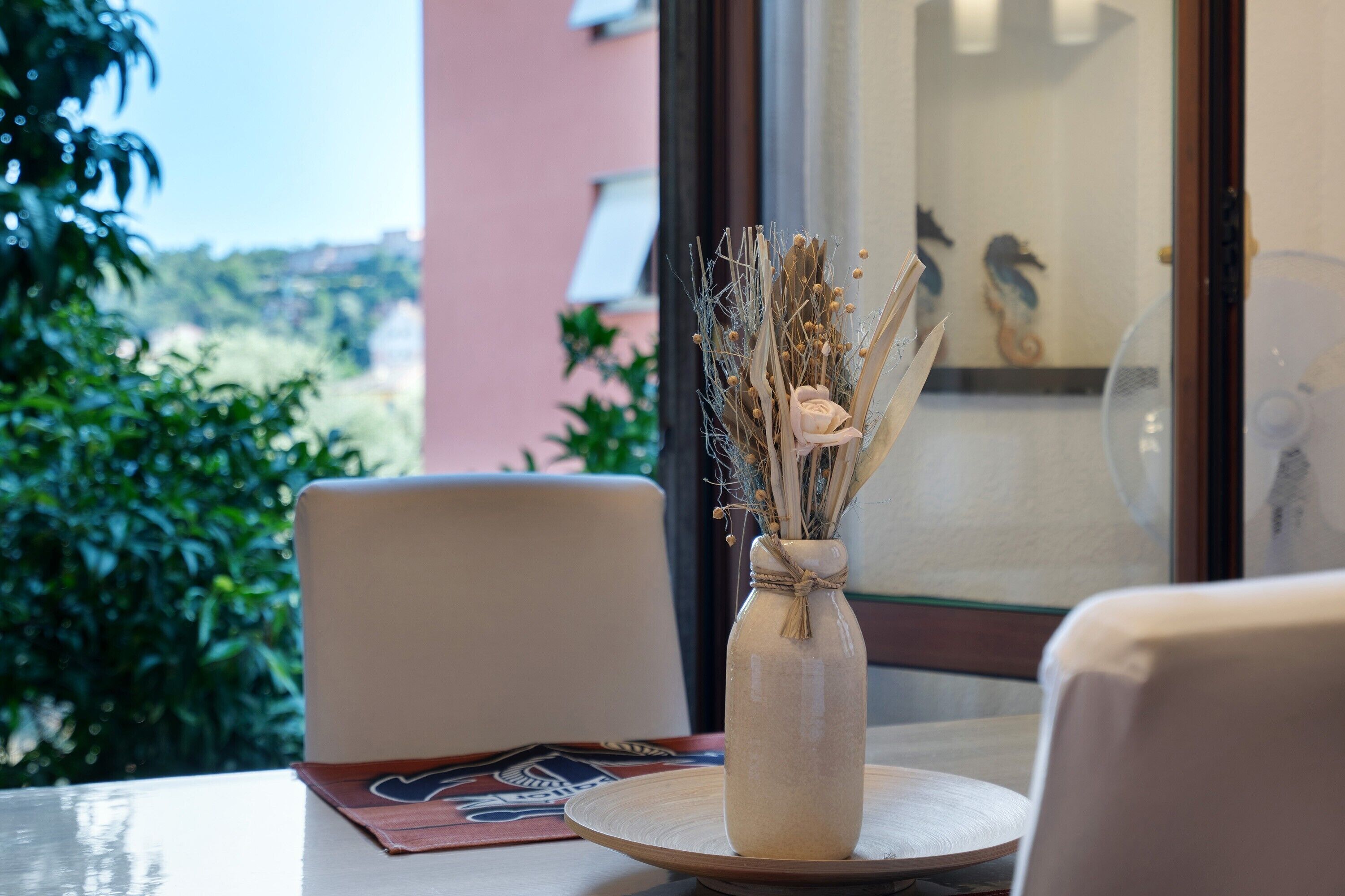 A table with a vase of dried flowers, a chair, and a window with a view of greenery.