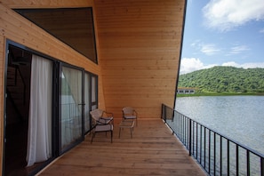 Een houten terras met uitzicht op een meer en heuvels, twee stoelen en een glazen deur.