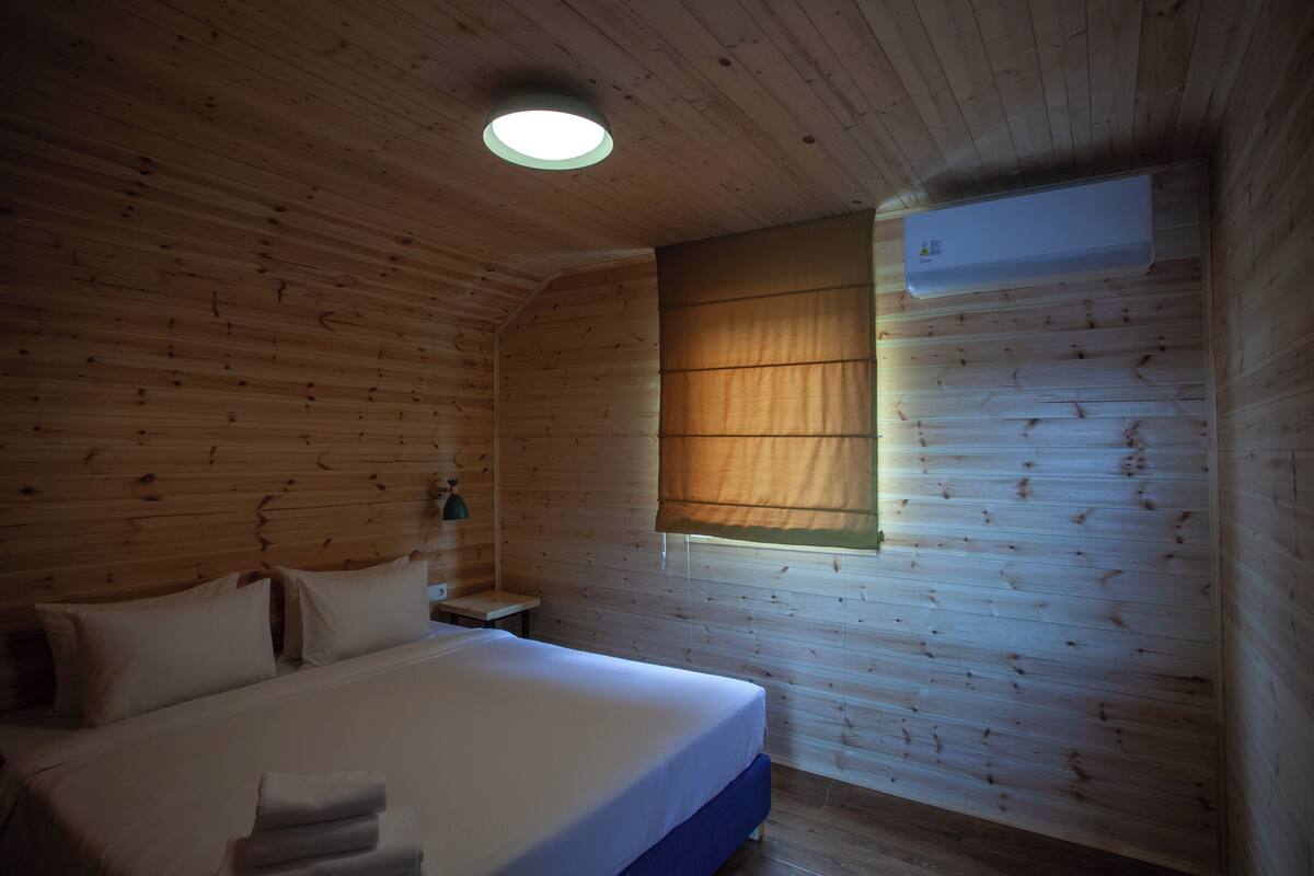 Een houten kamer met een bed, een nachtkastje, een lamp en een raam met jaloezieën.