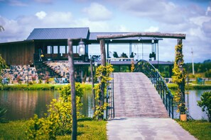 A wooden bridge leading to a building with a metal roof, surrounded by greenery and a body of water. - Moretoni Lake Resort  (Telavi)