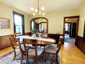 Une salle à manger avec une table en bois, des chaises, un lustre et un grand miroir.