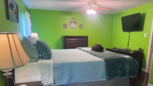 Un dormitorio con cama, mesita de noche, lámpara, ventilador de techo, televisor y pared verde.