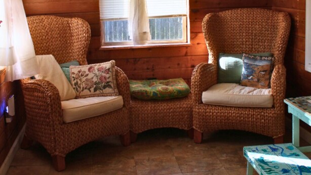 Una habitación acogedora con paredes de madera, dos sillas de mimbre con cojines, una mesita y una ventana con cortinas de encaje.