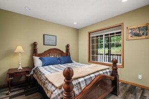 A bedroom with a wooden bed, a nightstand, a lamp, a window with a view of greenery, and a framed picture on the wall.