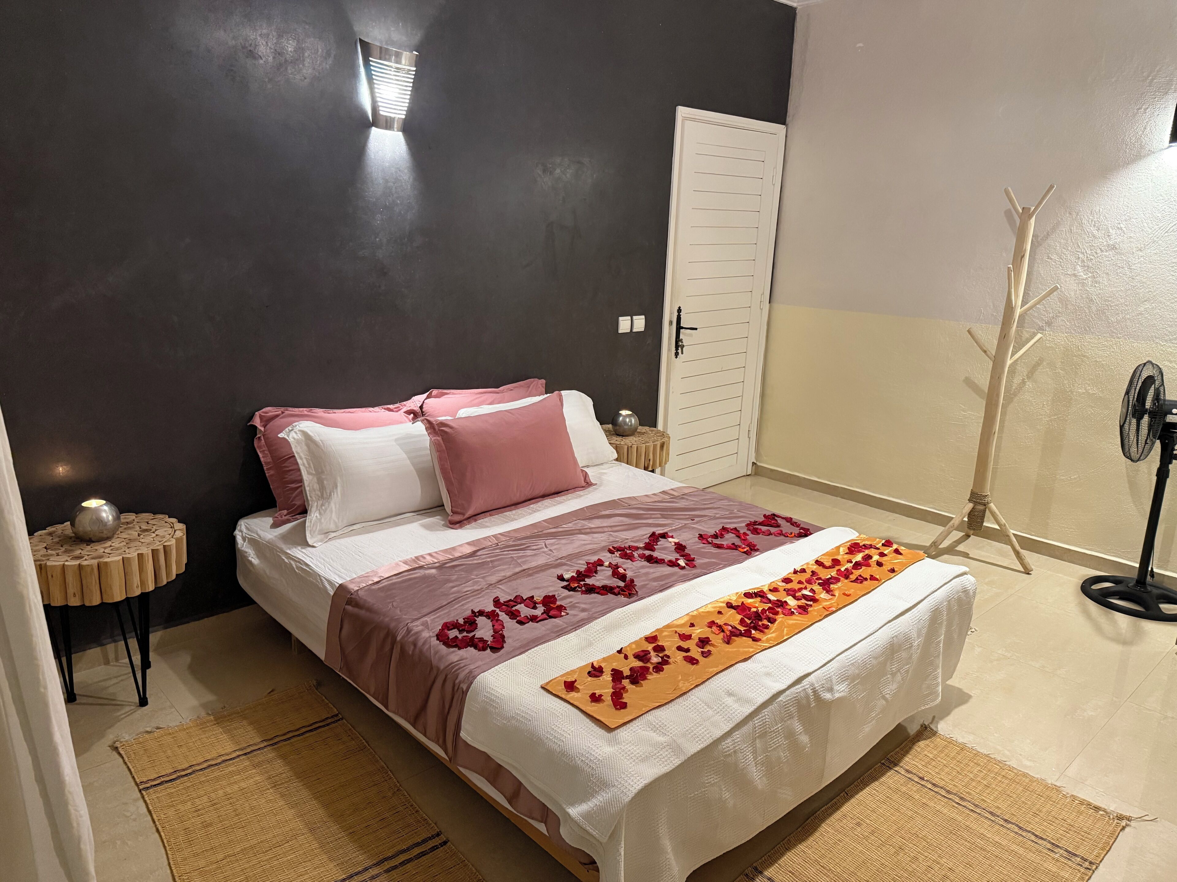 A neatly made bed with a white and pink bedspread, a decorative heart-shaped arrangement of rose petals, and a small table with a candle and a decorative bowl.