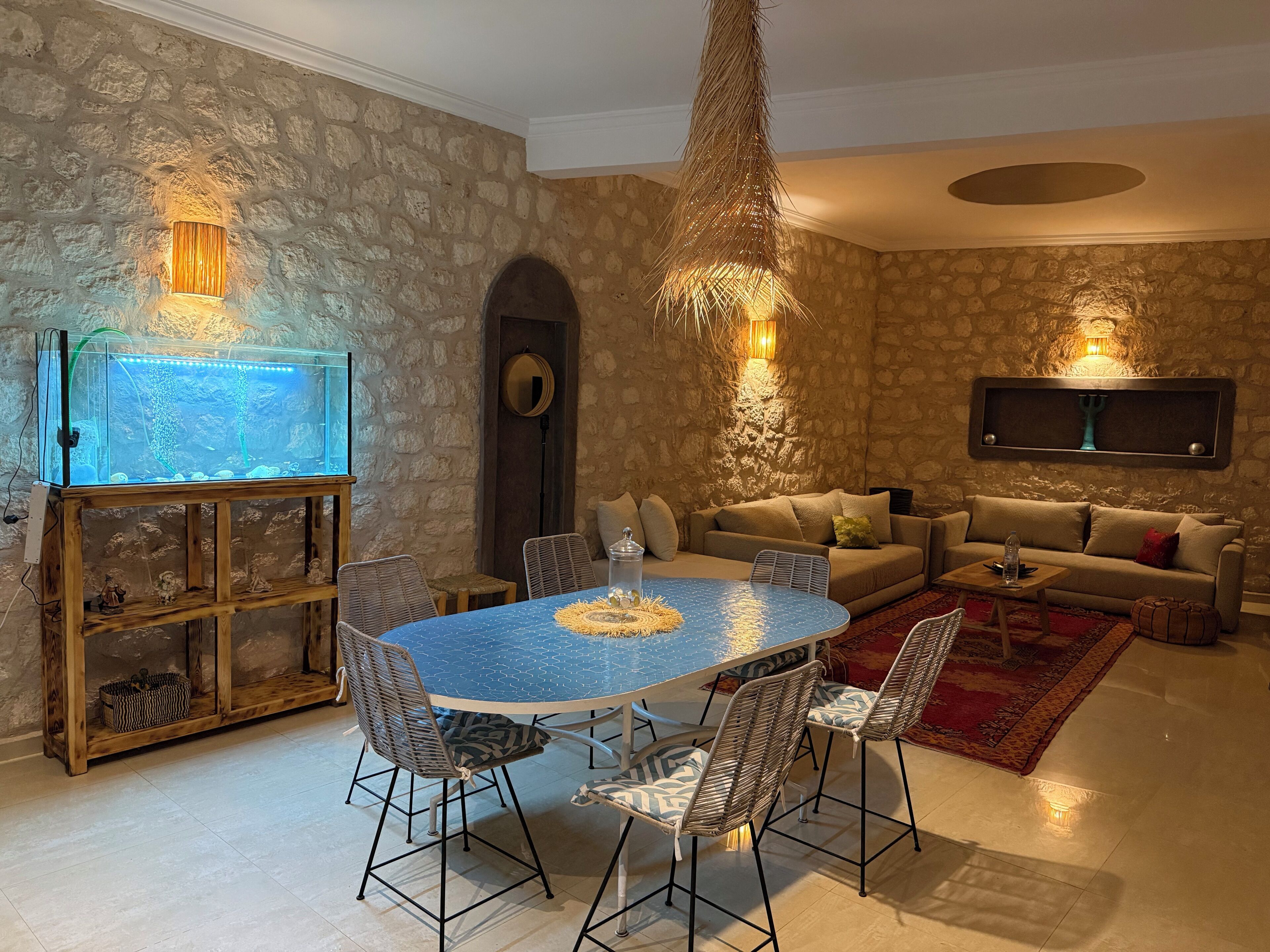 Una sala de estar acogedora con pared de piedra, una mesa redonda para comer, sillas de mimbre, un sofá y un televisor montado.