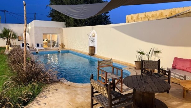 A pool area with a wooden table and chairs, a lounge chair, and a potted plant.