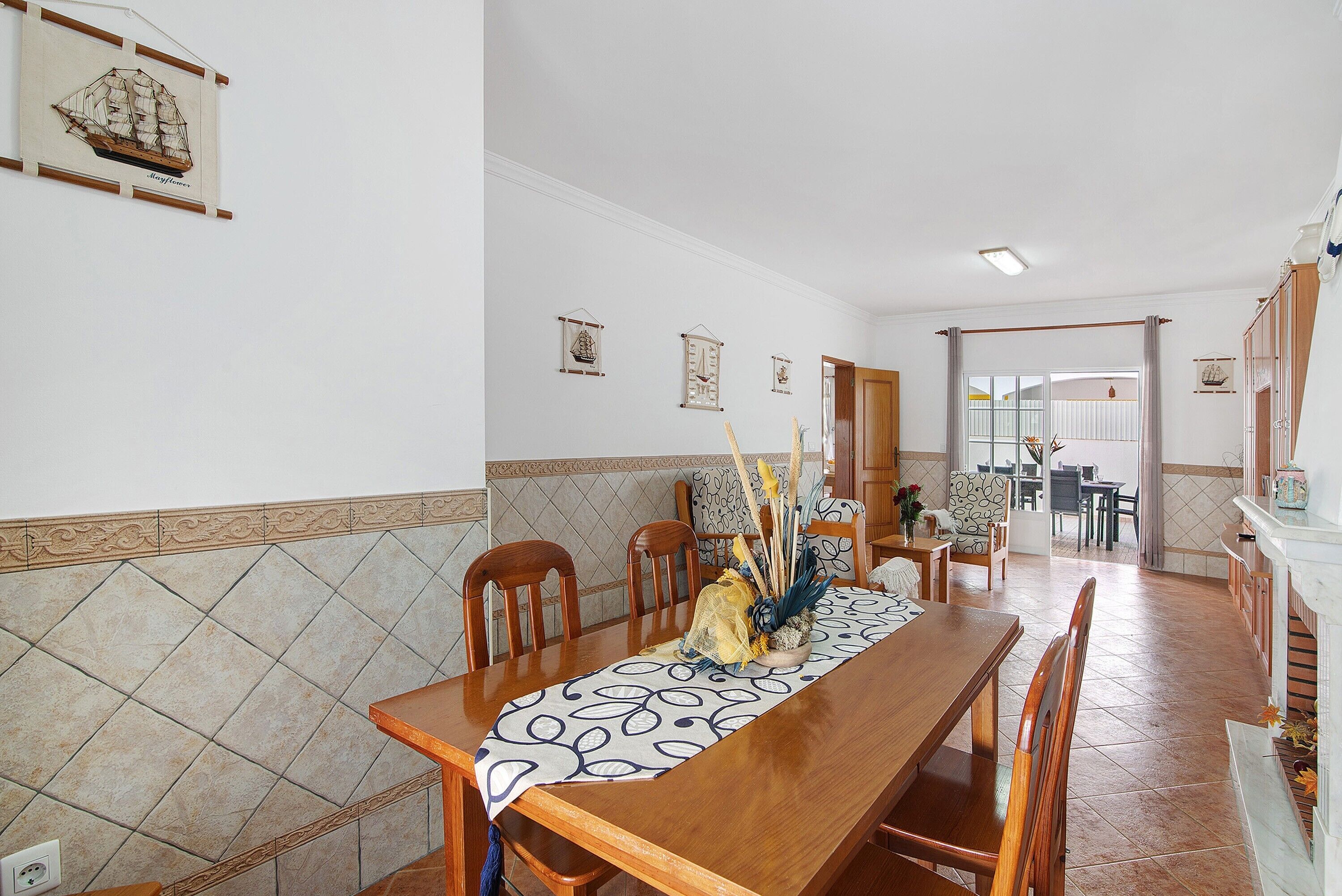 A dining area with a long wooden table, chairs, and a patterned tablecloth. There are wall-mounted decorations and a tiled backsplash.