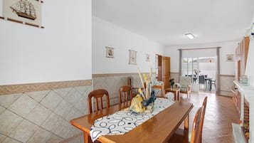 A dining area with a long wooden table, chairs, and a patterned tablecloth. There are wall-mounted decorations and a tiled backsplash.