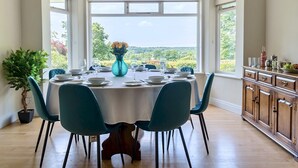 A dining area with a round table set for four, large windows offering a scenic view, and a wooden cabinet to the right. - 5bed Surrey home · Games Room · Near Thorpe Park - Pass the Keys (Surrey)