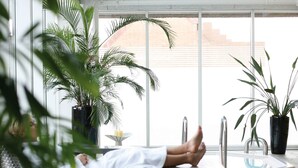 A person lying on a lounge chair in a well-lit room with large windows, surrounded by potted plants.