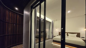 Une chambre d’hôtel moderne dotée d’une grande porte-fenêtre coulissante donnant sur un balcon, d’un lit soigneusement fait et d’une petite table de chevet.