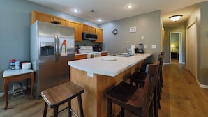 A modern kitchen with a stainless steel refrigerator, wooden cabinets, a microwave, and a sink. There are bar stools and a small table with a chair. - Quiet, Family Friendly 3/2 Home (Columbia)