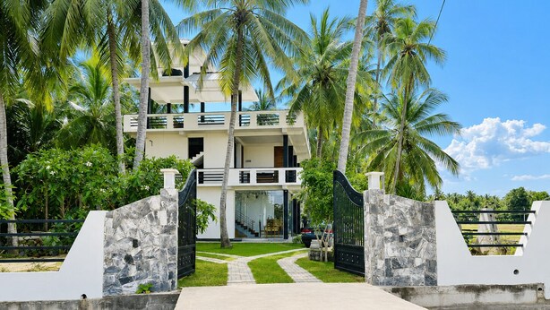 A modern multi-story building with a central staircase, surrounded by palm trees and a paved walkway leading to a gated entrance. - Tranquil Sanctuary Yala (Tissamaharama)