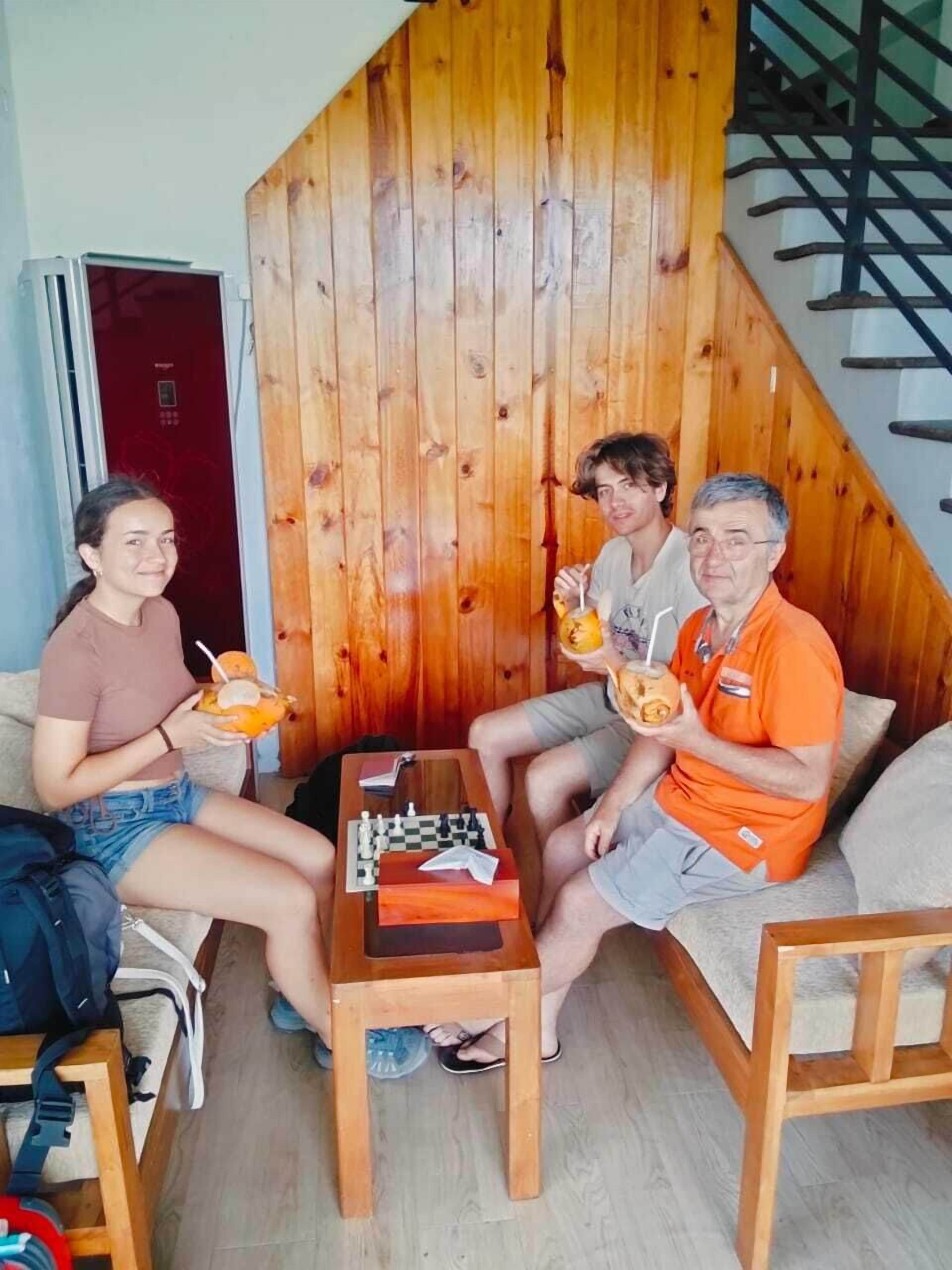 A cozy living room with wooden walls and a staircase. There is a chessboard on a small table, and three people are seated on a sofa, each holding a drink.