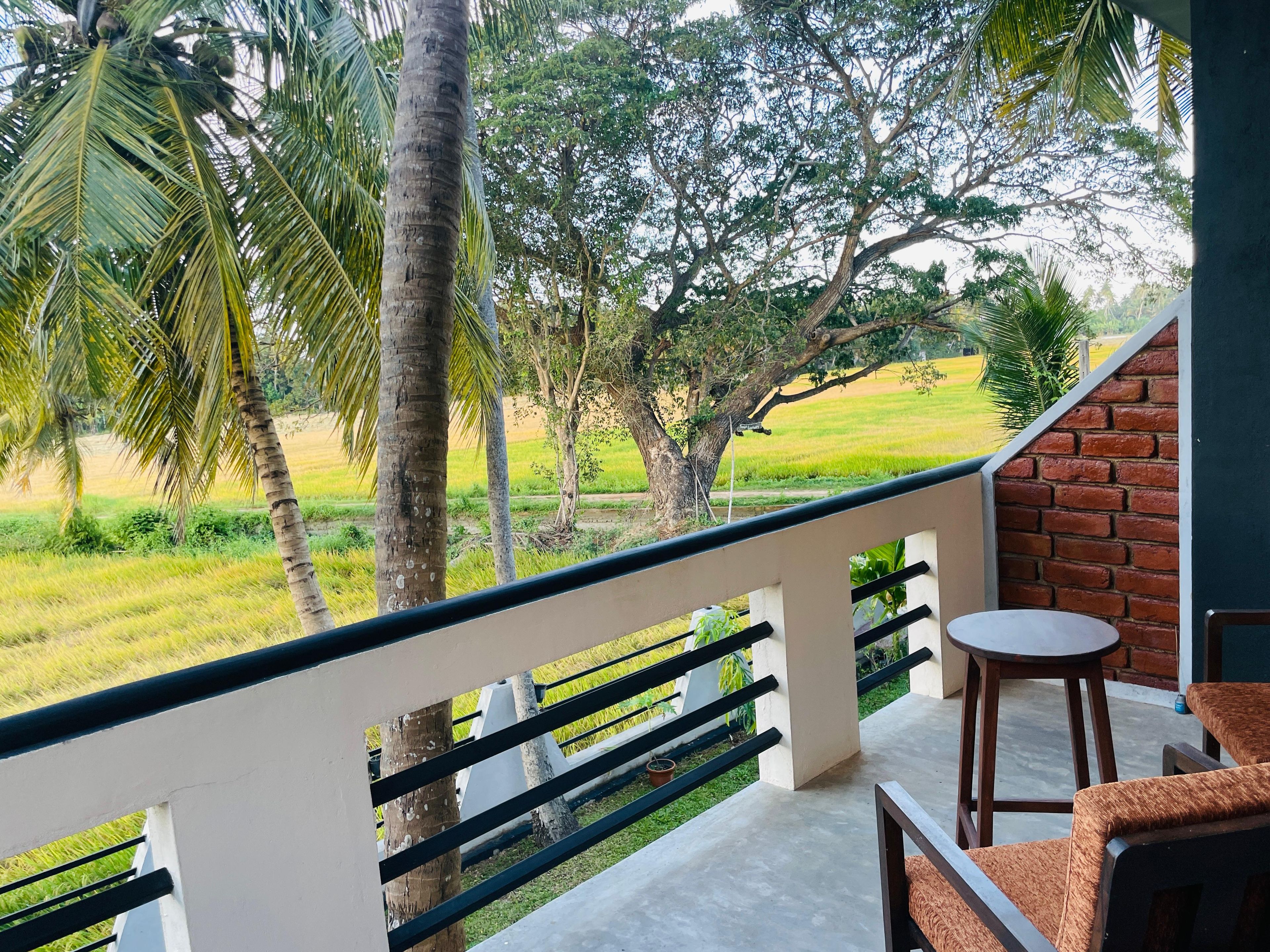 A balcony with a view of a green field and trees, a brick wall, and a small round table with a chair.