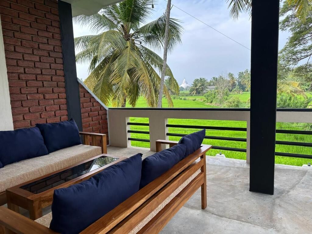 A covered outdoor seating area with a sofa and table, a brick wall, and a view of a green field and palm trees.