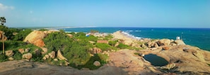 Un paysage cÎtier caractérisé par un terrain rocheux, une végétation luxuriante et une plage, avec des bùtiments visibles au loin.