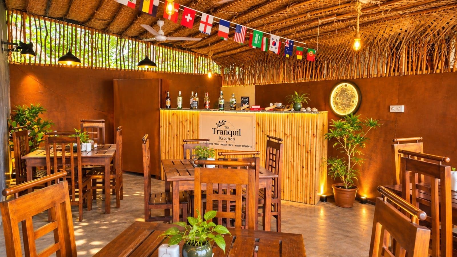 A restaurant interior with wooden tables and chairs, a bar area with a sign reading 'The Tranquil Kitchen,' and various flags hanging from the ceiling.