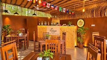 A restaurant interior with wooden tables and chairs, a bar area with a sign reading 'The Tranquil Kitchen,' and various flags hanging from the ceiling.