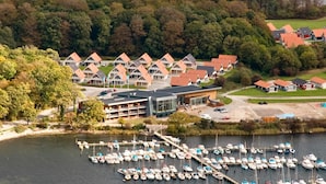 Un hotel frente al lago con varias construcciones, un puerto deportivo con numerosos barcos y una ladera boscosa al fondo.