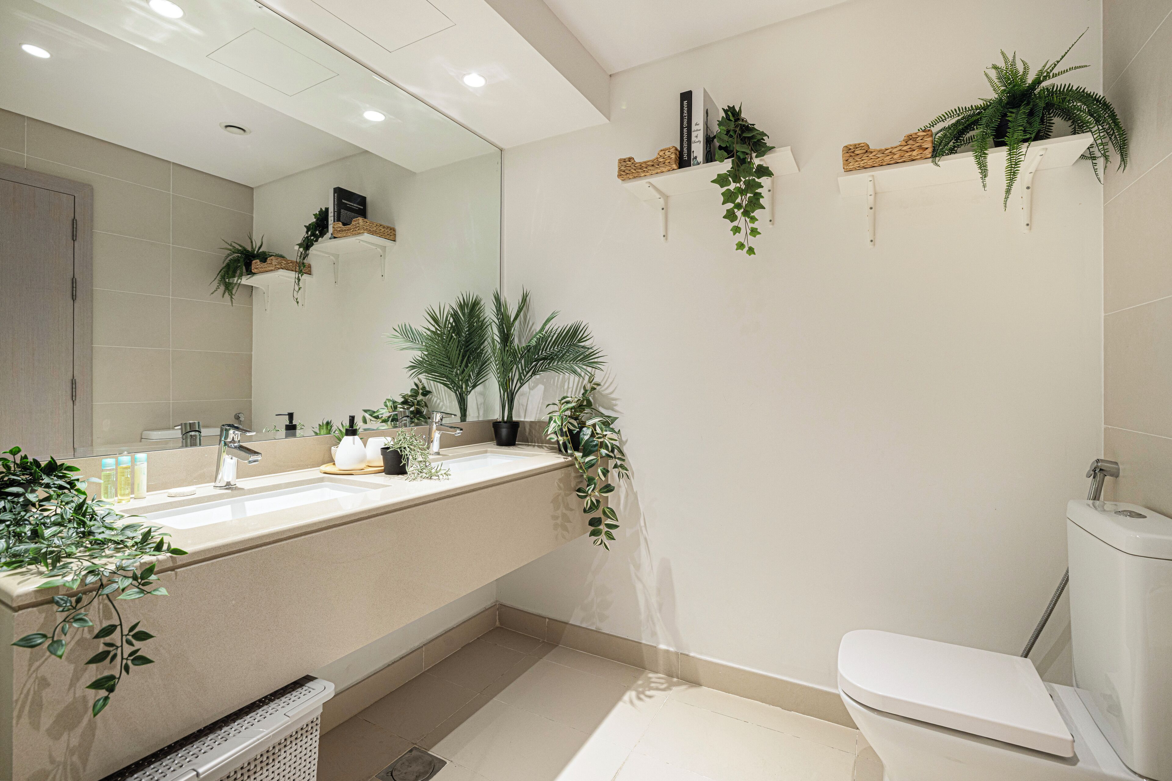 Un baño moderno con un espejo grande, un lavamanos y un inodoro. Hay estantes con plantas y artículos de aseo.