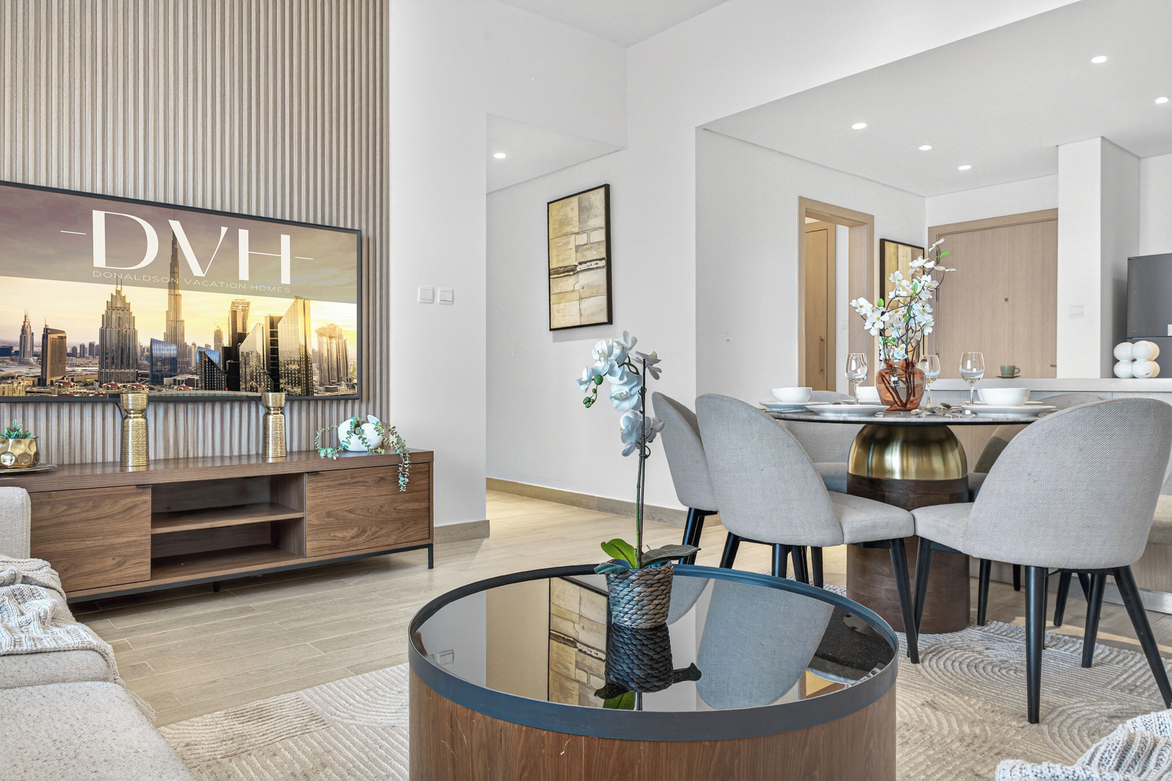 Una sala de estar moderna con una mesa de centro de vidrio, sillas grises, un mueble de entretenimiento de madera y una obra de arte con una vista urbana enmarcada.