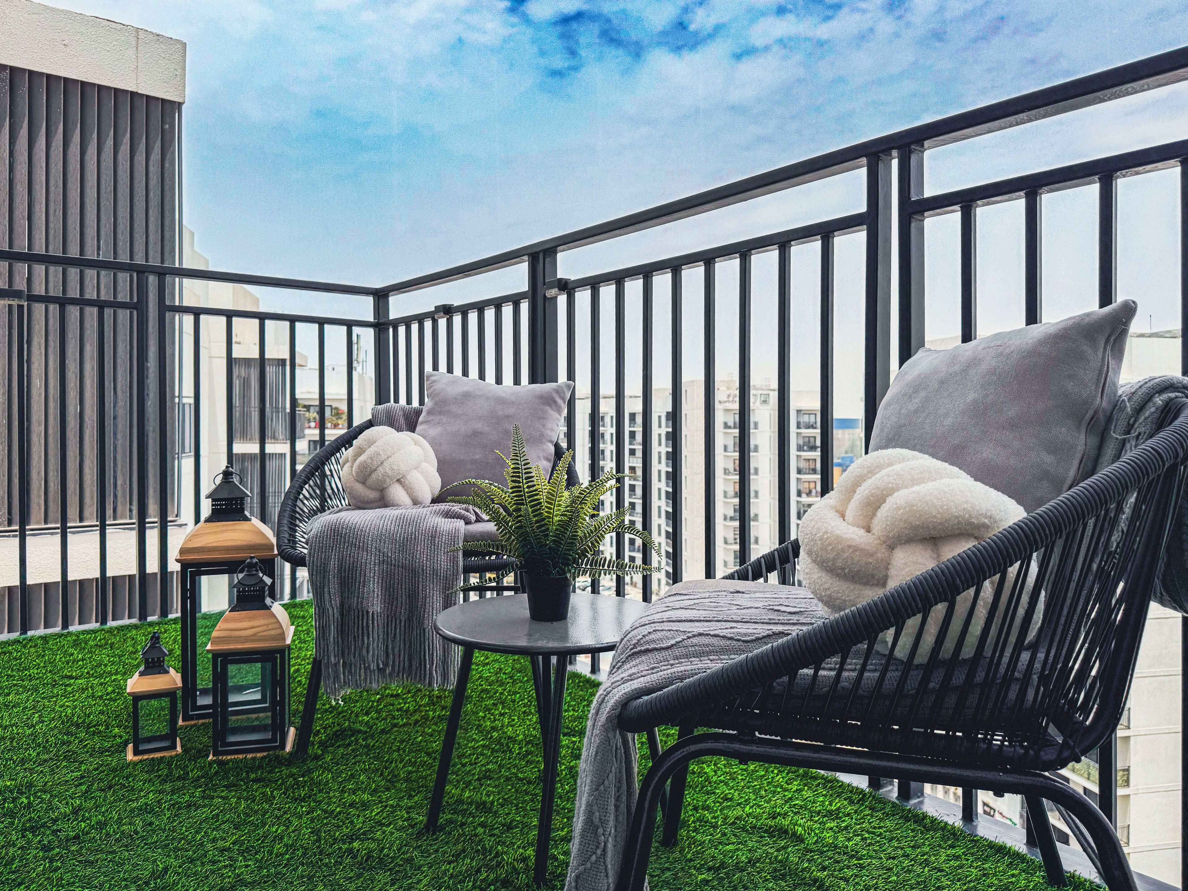 A balcony with a black metal railing, two wicker chairs with grey cushions, a small round table, and a potted plant.