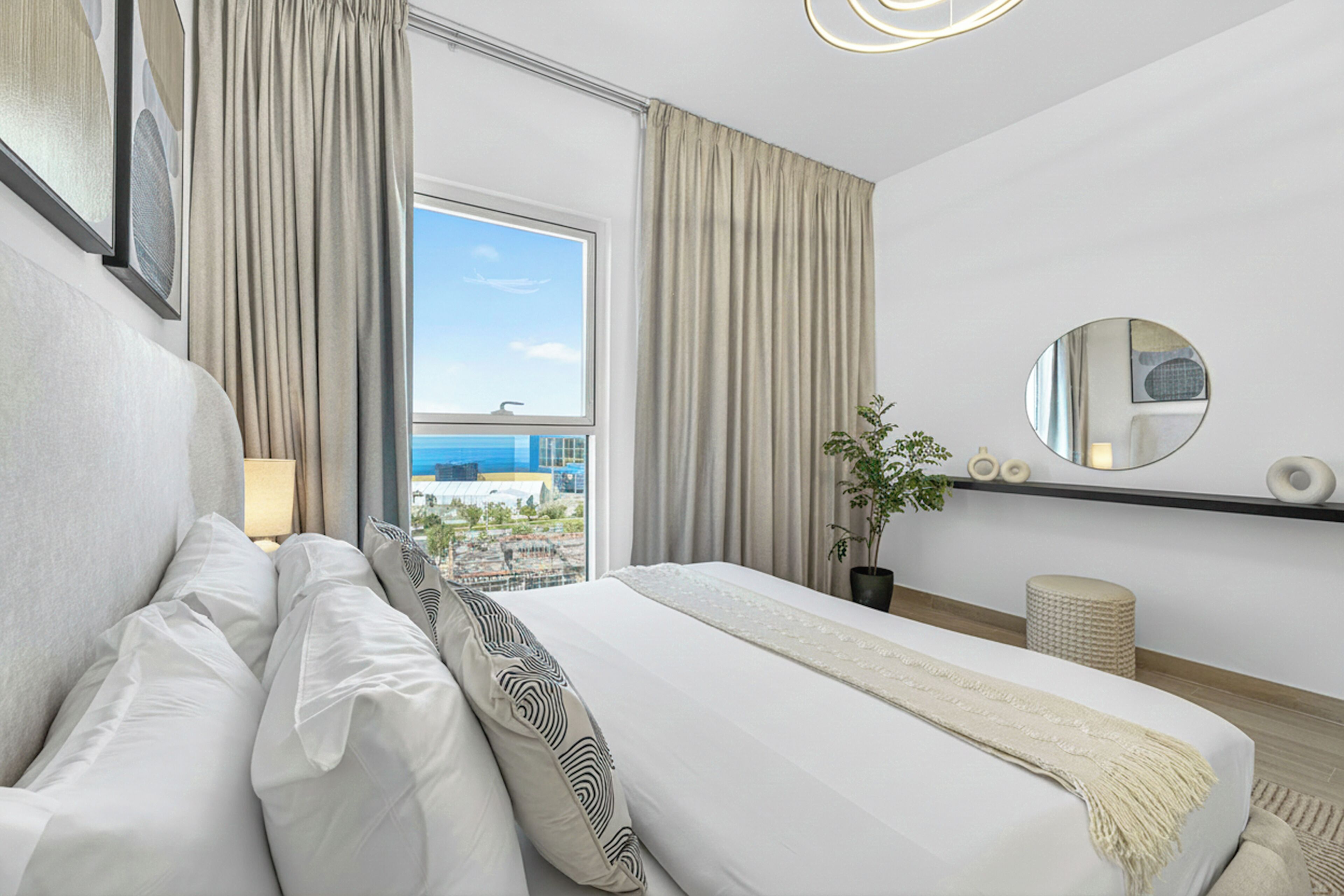 Ein Hotelzimmer mit einem Bett, einer Zimmerpflanze, einem runden Spiegel und Blick auf die Stadtlandschaft.