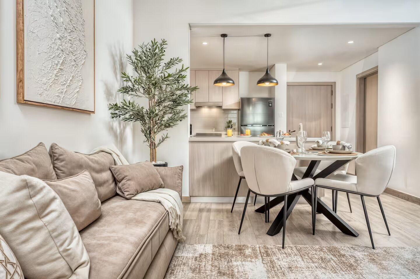 A modern living room with a beige sofa, a dining table with chairs, a kitchen area with pendant lights, and a large plant.