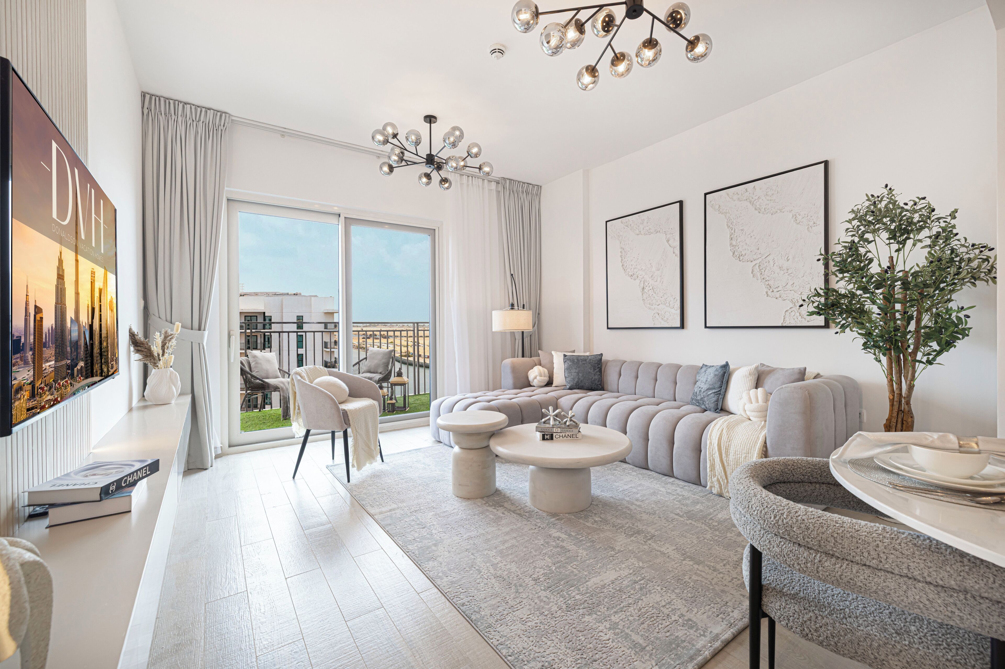 A modern living room with a large sofa, a coffee table, and a dining area with a round table. There is a large window with curtains, a chandelier, and framed artwork on the wall.
