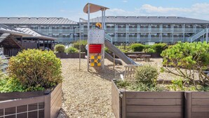 A playground with a slide and climbing structures, surrounded by wooden fences and greenery, with a modern building in the background. - 4 Person Holiday Home on a Holiday Park in Fano (Fano)