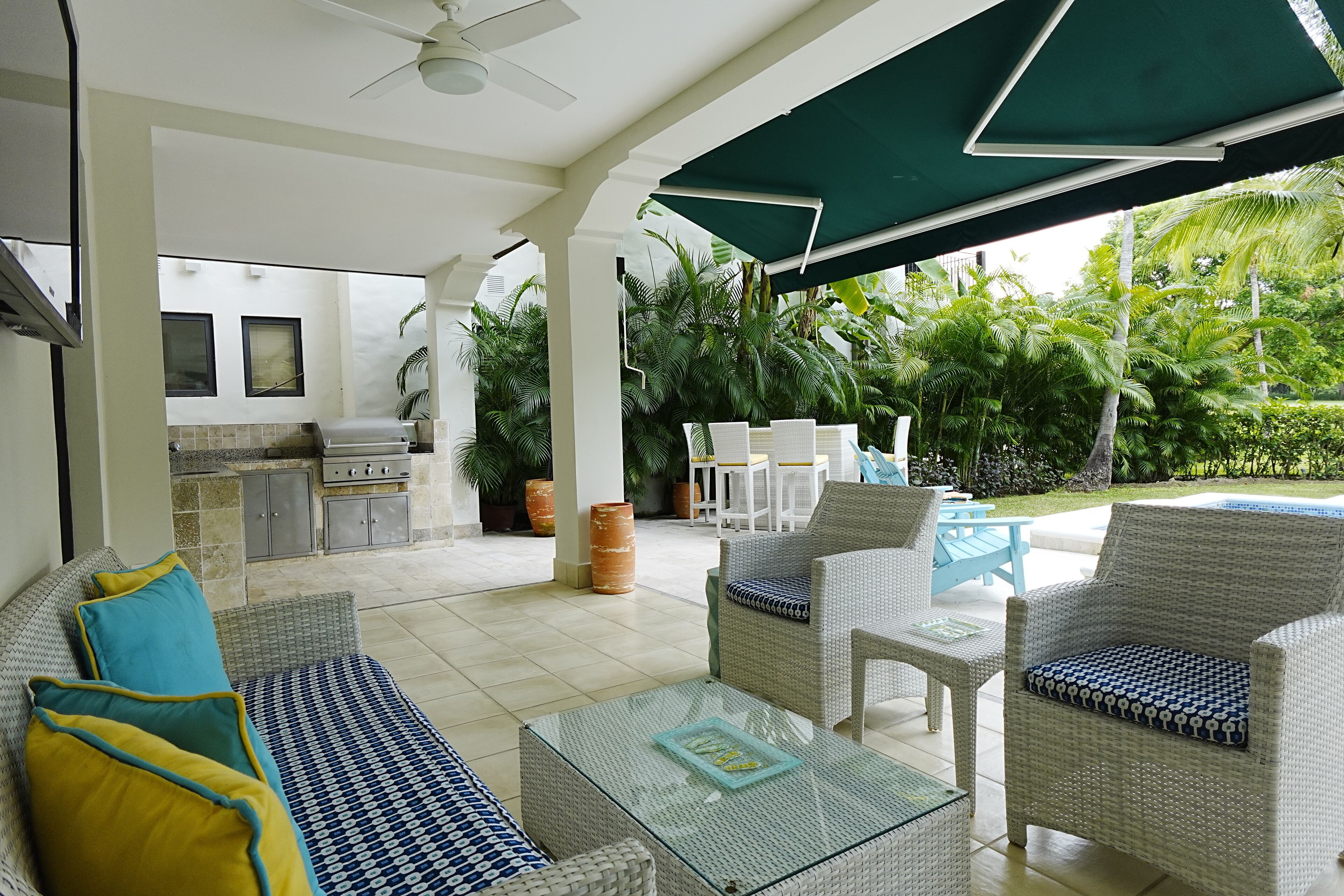 Un área de estar al aire libre techada, con mobiliario de mimbre, una mesa de vidrio y vistas a una piscina y vegetación.