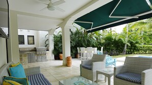 A covered outdoor seating area with wicker furniture, a glass table, and a view of a pool and greenery.