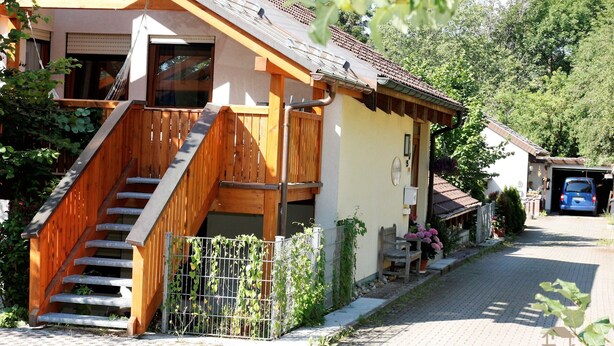 Un bâtiment à deux étages avec un escalier en bois menant à un balcon, une véranda et un véhicule garé dans l’allée.