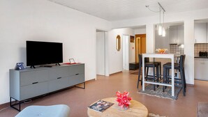 Un salon moderne doté d’un téléviseur à écran plat, d’un meuble de rangement gris, d’une table basse ronde avec des magazines, et d’un coin repas comprenant une table blanche et des chaises noires.