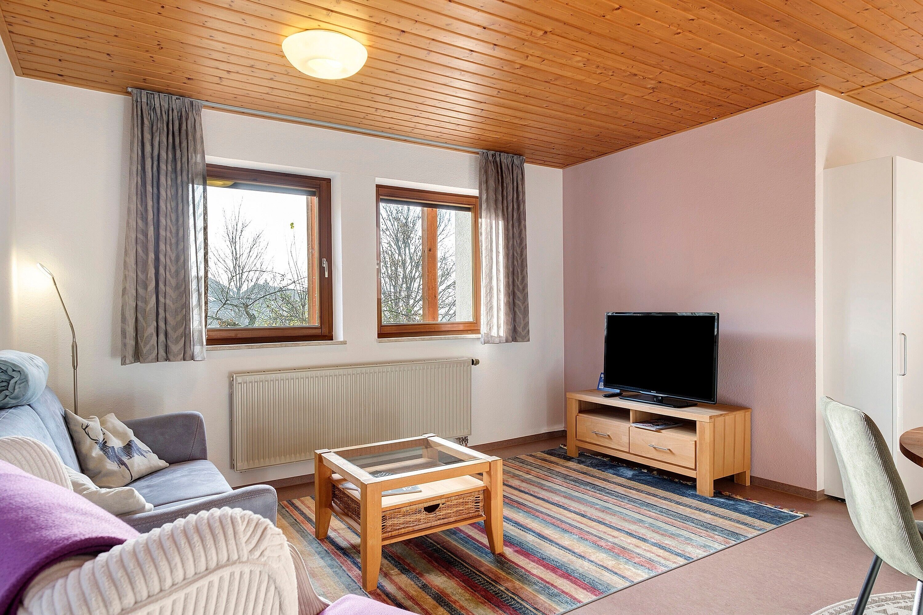 A living room with a sofa, a coffee table, a TV on a wooden stand, and a window with curtains.