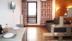 Una habitación con una mesa de comedor preparada para dos personas, un sofá, un televisor montado en la pared y un ventanal con cortinas estampadas.