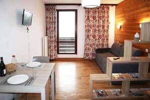 A room with a dining table set for two, a sofa, a TV mounted on the wall, and a window with patterned curtains.