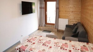 Ein Zimmer mit Holzeinrichtung, einer Couch, einem Tisch mit gemustertem Tuch und einem an der Wand befestigten Fernseher, sowie einer mit Vorhängen versehenen Tür.