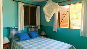 A bedroom with a bed, two pillows, a nightstand, a lamp, a window with curtains, and a view of the outside. - Sea view bungalow (Bouillante)