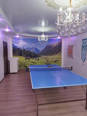 A room with a ping pong table, a chandelier, and a mural of a mountainous landscape. - Travel Around Guest House (Kochkor)