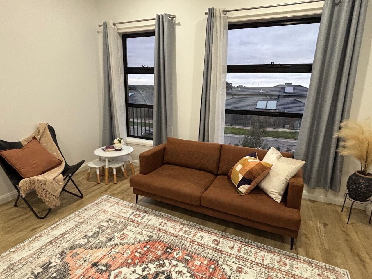 A living room with a brown sofa, a patterned rug, a small round table, and two windows with curtains.