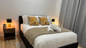 Ein ordentlich gemachtes Bett mit einer weiß-beigen Decke, zwei Kissen mit geloben-Blatt-Muster und einem gefalteten Handtuch. Zwei Nachttische mit Lampen, ein dunkles Kopfteil und ein grauer Vorhang am Fenster sind ebenfalls zu sehen.