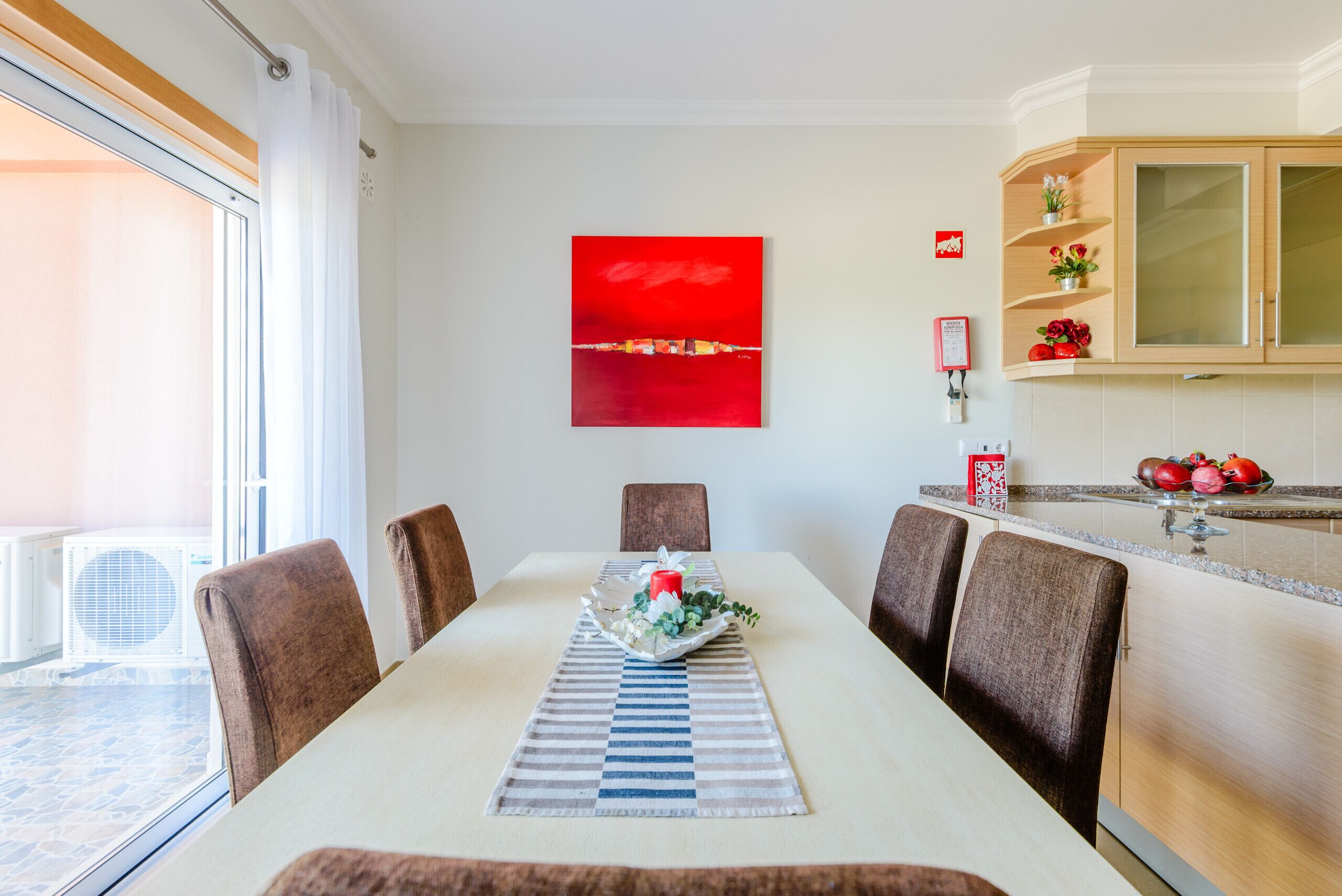 A dining area with a table set for four, a red abstract painting on the wall, and a kitchen with wooden cabinets and a fruit bowl on the counter.
