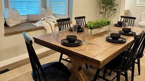 Un coin repas avec une table en bois, des chaises noires et une fenêtre équipée de stores.