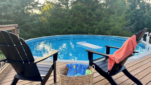 Une terrasse en bois avec deux chaises Adirondack noires, donnant sur une piscine, le tout entouré d’arbres.