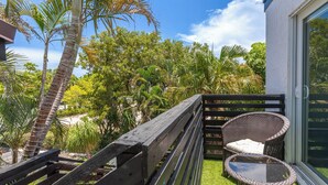 A balcony with a wicker chair and a small table, surrounded by lush greenery and a clear blue sky.